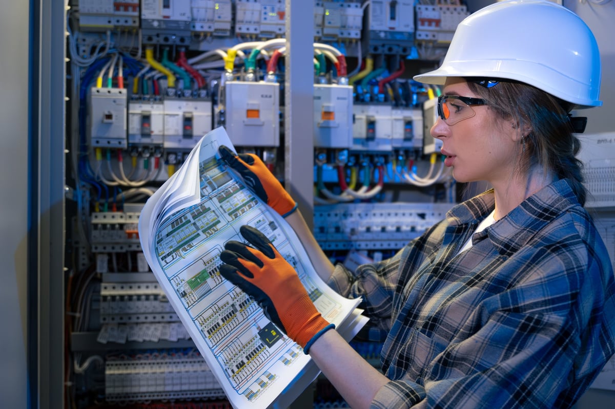 A woman electrician in gloves points at a blueprint while validating a cabinet node.
