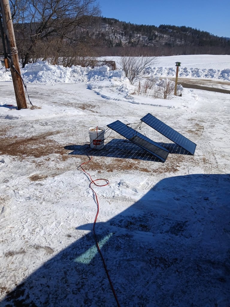 Portable solar panels on snowy ground by a frozen lake with bare trees and mountains in background