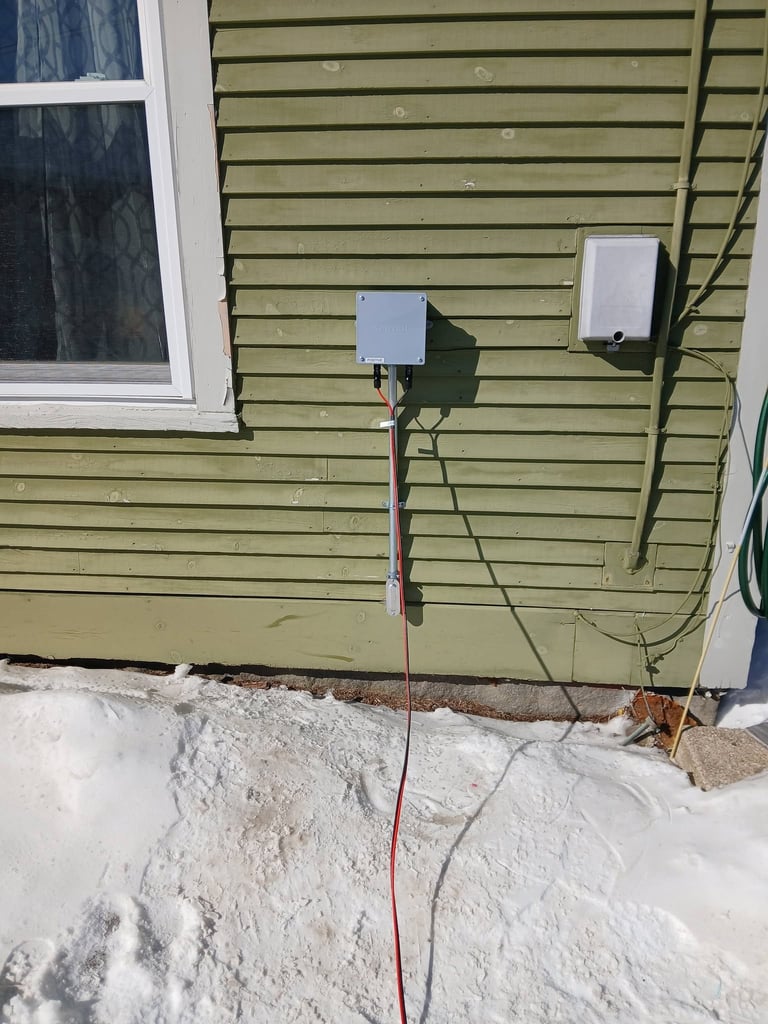 Electrical boxes mounted on green horizontal siding with cables running down to snowy ground and wall base