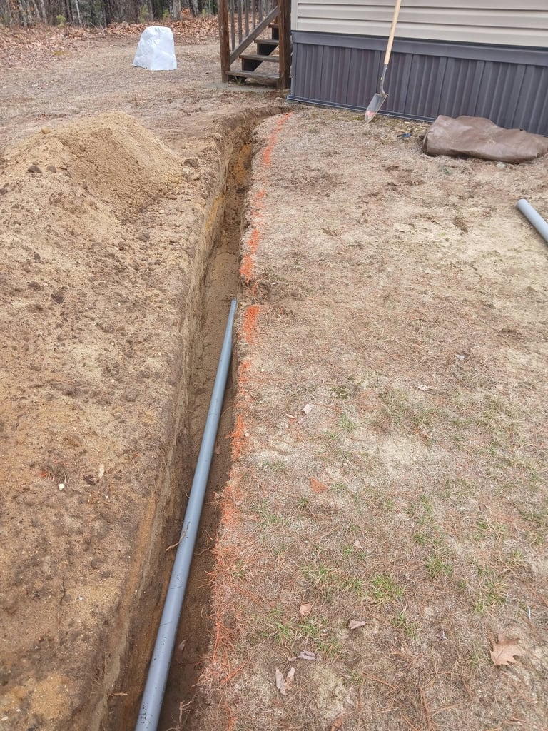 A long trench dug in bare ground with a gray PVC pipe running through it, near a metal building with landscaping materials visible