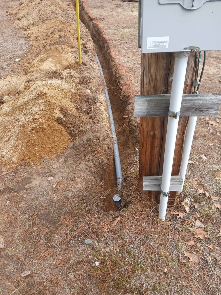 Underground utility lines and pipes including white PVC and gray conduit pipes installed in a trench next to excavated soil