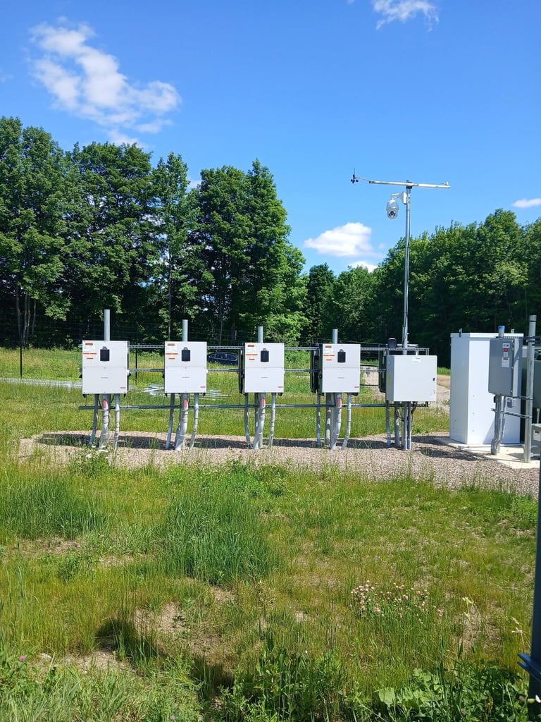 Row of white weather monitoring stations in a grassy field with trees and a measurement tower against a blue sky