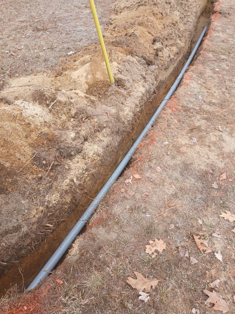 Excavated trench with gray pipe and yellow measurement stick in soil during construction work